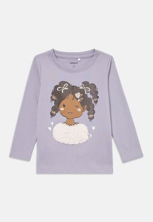 Lilaksi värjätty pitkähihainen paita, jossa on sarjakuvatyylinen tyttökuvitus. Tyttö on tummatukkaisena, hänen hiuksissaan on nutturoita ja rusetteja, ja hänellä on valkoinen teksturoitu yläosa. Kuvan ympärillä on pieniä sydämiä.