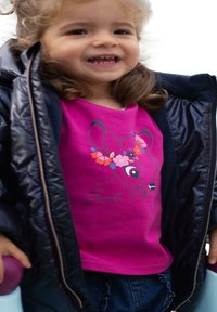 Un enfant souriant portant un t-shirt Bambi rose avec des fleurs, une veste doudoune noire et tenant un objet violet.