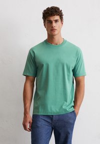 Zielony bawełniany t-shirt z okrągłym dekoltem i krótkim rękawem, charakteryzujący się gładką fakturą i luźnym krojem, w zestawieniu z ciemnoniebieskimi dżinsami.