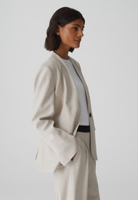 Beige blazer met één knoop en aangeknipte revers. Gemaakt van gestructureerde stof, heeft brede manchetten en zijzakken. Gecombineerd met een wit topje.