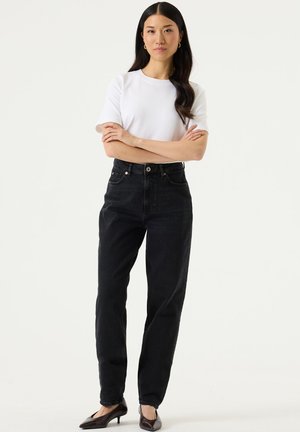 Femme debout, bras croisés, portant une chemise blanche à manches courtes, un jean noir taille haute et des chaussures marron à talons bas, sur fond uni.