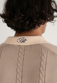 Sweter dzianinowy w odcieniu taupe z teksturowanym wzorem w warkocze, z kremowym ściągaczem przy kołnierzu i haftowanym logo na plecach.