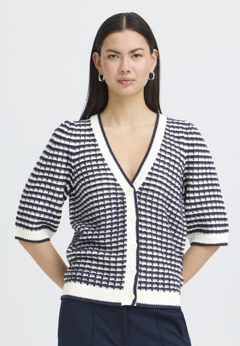 Femeie cu păr drept, închis la culoare, poartă un cardigan cu nasturi în carouri bleumarin și alb, cu mâneci până la cot, stând în picioare în fața unui fundal simplu.