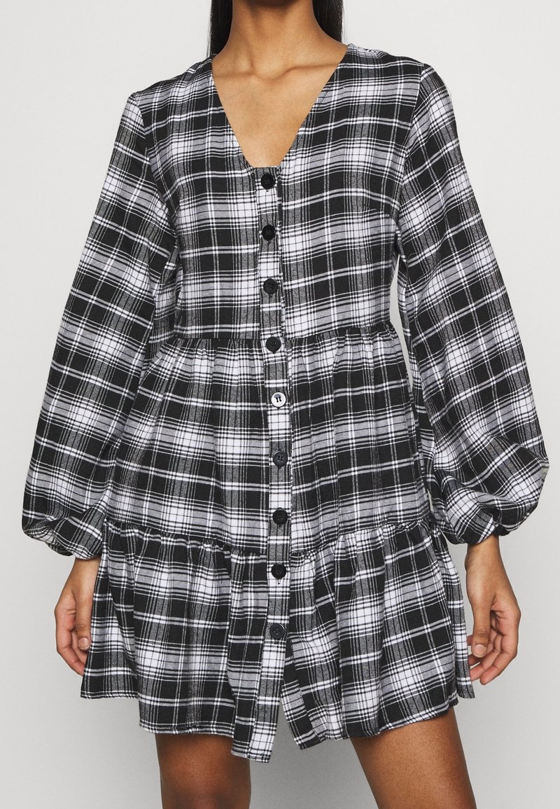 Femme portant une robe chemise à carreaux noirs et blancs avec de longues manches bouffantes et un décolleté en V, debout avec les mains détendues sur les côtés.
