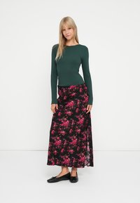Lang sort nederdel med pink blomsterdesign, parret med en tætsiddende grøn langærmet top. Modellen har sorte flade sko på.