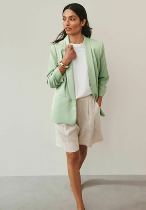 Kobieta o długich ciemnych włosach, ubrana w jasny zielony blezer, białą koszulę, beżowe szorty i złotą bransoletkę, stoi na tle jednolitego jasnego tła.