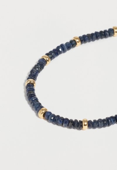 Βραχιόλι με πολύεδρες σκούρες μπλε χάντρες διακοσμημένες με μικρές, λεία χρυσά διαχωριστικά χάντρες σε απλό ανοιχτό φόντο.