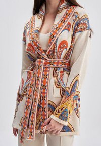 Cardigan en tissu tissé avec une base beige, présentant des motifs paisley orange et bleu complexes, une ceinture et des poches latérales. Texture douce et coupe décontractée.