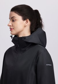 Mujer con cabello oscuro recogido en una coleta, vistiendo una chaqueta impermeable negra con capucha y el logo "ICEPEAK" en la manga, mirando hacia la izquierda.