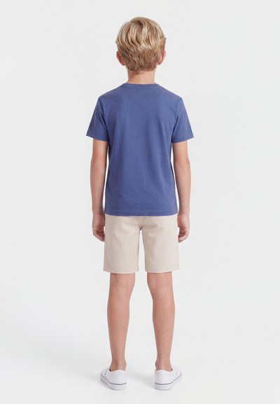 Ξανθό αγόρι που στέκεται με το πίσω μέρος στραμμένο, φοράει μπλε κοντομάνικο πουκάμισο, μπεζ σορτς και λευκά αθλητικά παπούτσια σε λευκό απλό φόντο.