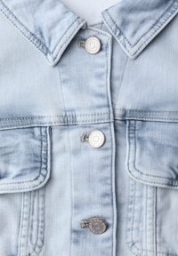 Lyseblå denimjakke med metaltryklåseknapper, brystlommer med klapper, krave samt synlige sømme og stikningsdetaljer.