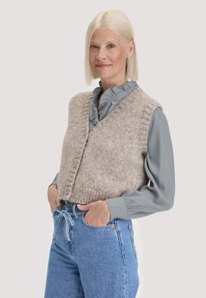Woman with straight blonde hair wears a gray ruffled blouse, beige knitted vest, and blue jeans with hands in pockets, standing against plain background.