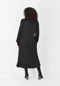 Robe noire longue avec des manches longues, texture lisse, ourlet légèrement évasé et manches élastiquées, associée à des bottes noires à talons arrivant à la cheville.