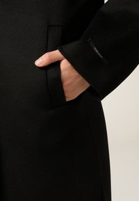 Cappotto nero in lana con una consistenza liscia, tasche laterali e un'etichetta discreta sulla manica. Design su misura con una vestibilità rilassata.