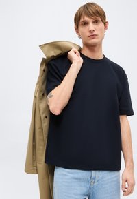 Νέος άνδρας με μαύρο κοντομάνικο πουκάμισο με υφή και ανοιχτό μπλε τζιν, κρατώντας μπεζ παλτό στον ώμο, στέκεται μπροστά σε απλό φόντο.