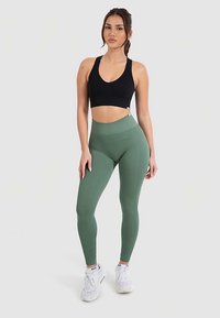 Sujetador deportivo negro y leggings verdes acanalados, de cintura alta. Los leggings tienen un patrón texturizado. La modelo lleva zapatillas deportivas blancas.