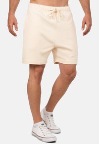Beige katoenen shorts met een elastische tailleband en trekkoord; voorzien van zijzakken en subtiele stikseldetails. Draagt met witte sneakers.