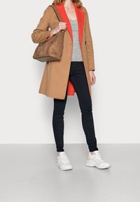 Abrigo marrón claro con cuello rojo, camiseta sin mangas gris, jeans oscuros y zapatillas blancas. Lleva una bolsa marrón. Texturas suaves y diseño moderno.