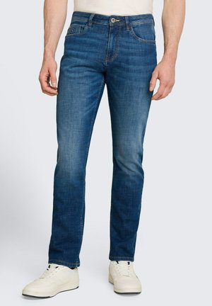 Mann trägt Slim-Fit blaue Jeans und weiße Sneakers, steht vor einem einfarbigen hellgrauen Hintergrund.