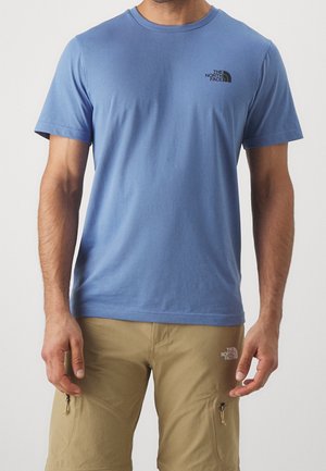Homme portant un T-shirt bleu clair à manches courtes et un short cargo marron clair, tous deux avec le logo The North Face sur le côté gauche.