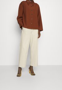 Brązowa koszula na guziki, kremowe spodnie o szerokich nogawkach oraz wzorzyste szpilki z żółtymi akcentami. Kluczowe cechy: luźny krój, fakturowana tkanina.