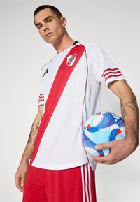 Vitt och rött sporttröja med diagonal rand, tillsammans med röda shorts. Spelaren håller en blå fotboll. Har laglogotypen på bröstet.