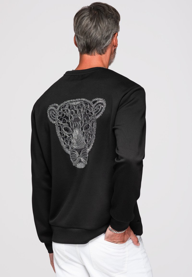 Homme aux cheveux gris portant un sweat-shirt noir avec une broderie abstraite argentée représentant un visage d'animal dans le dos et un pantalon blanc.