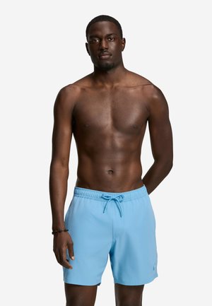 Hombre sin camisa de pie frente a un fondo blanco, vistiendo pantalones cortos de baño azul claro con cordón, mirando hacia adelante con expresión neutral.