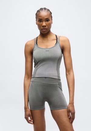 Donna con capelli intrecciati che indossa un top sportivo grigio senza maniche e pantaloncini abbinati a vita alta, in piedi contro uno sfondo chiaro e semplice.