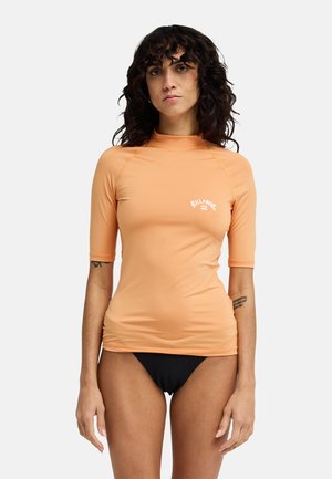 Kobieta o kręconych ciemnych włosach, ubrana w pomarańczowy krótkim rękawem rash guard z logo "Billabong" i czarne bikini, stojąca na białym tle.