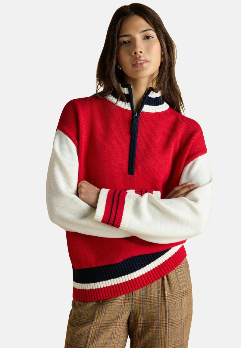 Rot-weißer Strickpullover mit hohem Kragen, ausgestattet mit einem Reißverschluss und marineblauen Streifenakzenten an den Ärmeln und dem Saum. Strukturiertes Gewebe.