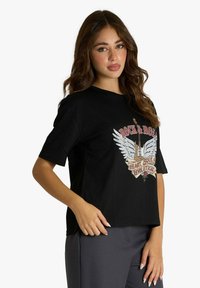 Maglietta nera in cotone con maniche corte, caratterizzata da un design grafico con ali e una chitarra in argento e accenti colorati.