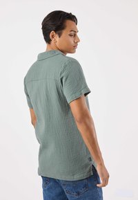 Camicia a maniche corte in tessuto verde salvia testurizzato, con scollo con colletto e design con spacco sul fondo. Modello visto da dietro.