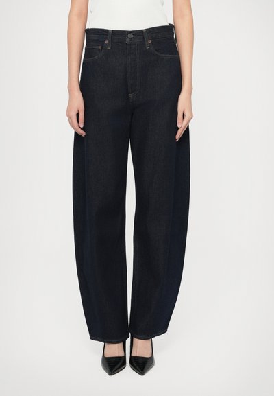 Jean denim foncé classique à jambe large et taille haute, doté de poches avant, d'une fermeture par boutons et de coutures nettes. Associé à des talons pointus noirs.