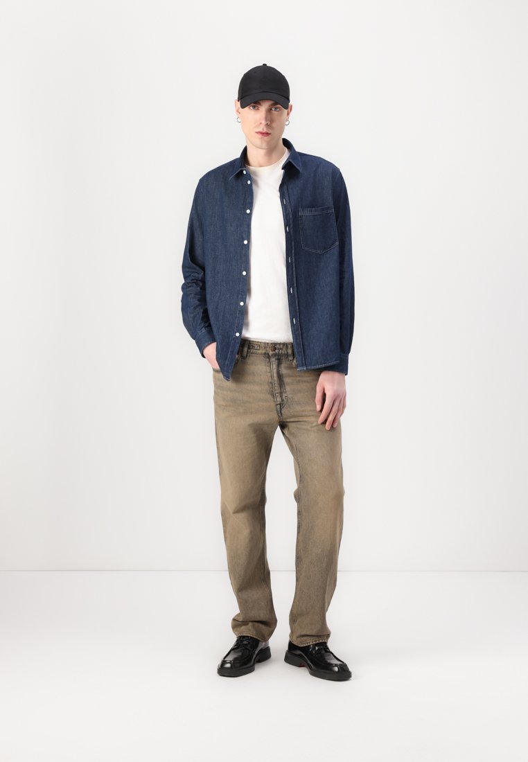 Chemise en denim bleu foncé, t-shirt blanc, pantalon marron clair et chaussures noires. Le mannequin porte une casquette noire et se tient devant un fond blanc.
