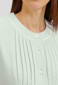 Top de punto verde claro con un patrón en forma de zigzag, que presenta pliegues, un cuello redondo y cierres de botón. Los acentos incluyen ribete blanco.