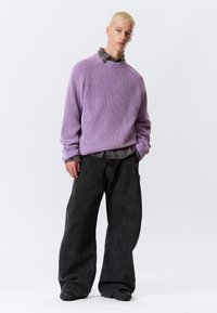 Pull en tricot lavande surdimensionné avec texture côtelée porté sur une chemise grise à boutons, associé à un pantalon en denim noir ample et des chaussures noires.