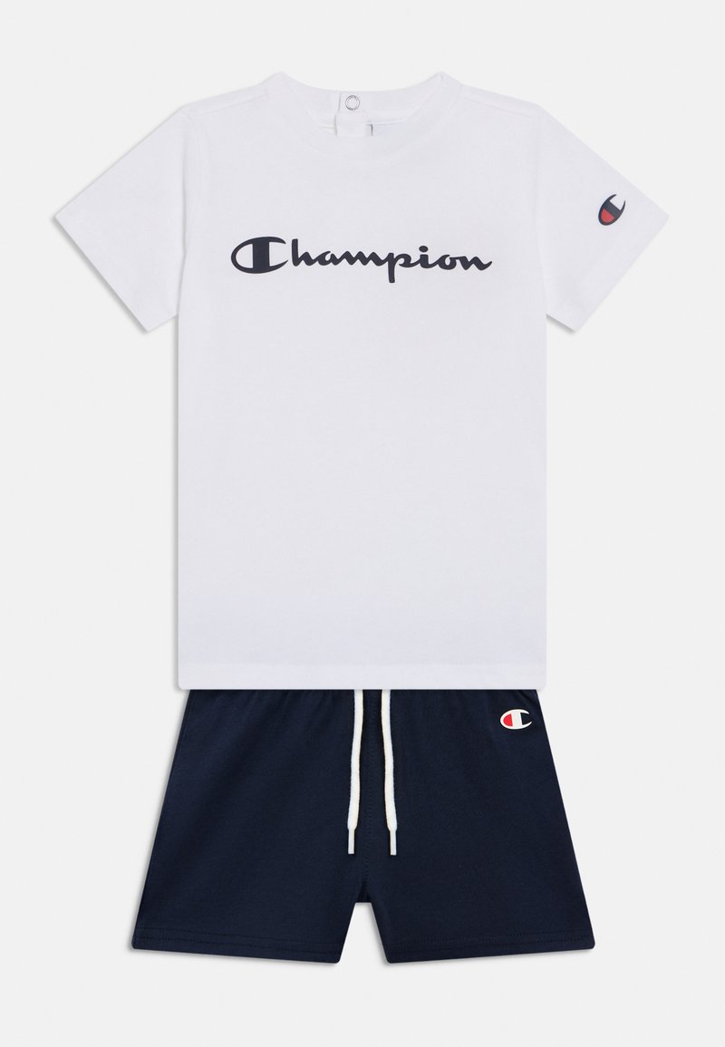 Λευκό βαμβακερό μπλουζάκι με μαύρο λογότυπο «Champion», συνδυασμένο με σκούρο μπλε σορτς που έχει κορδόνι στη μέση και μικρό ταμπελάκι λογότυπου στο πόδι.