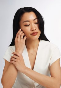 Femme aux longs cheveux noirs portant un haut blanc en V, des bagues en or, et un délicat collier en or avec deux cercles entrelacés, regardant vers le bas pensivement.