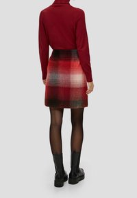 Femme portant un col roulé rouge, une jupe écossaise rouge, des collants noirs transparent et des bottines noires se tenant devant un fond uni.