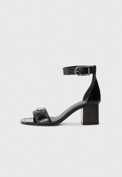 Sandal cu toc înalt din piele neagră, cu vârf pătrat și brățară pentru gleznă. Prezintă o bandă frontală plată și un accent cu cataramă metalică. Design cu toc pătrat.