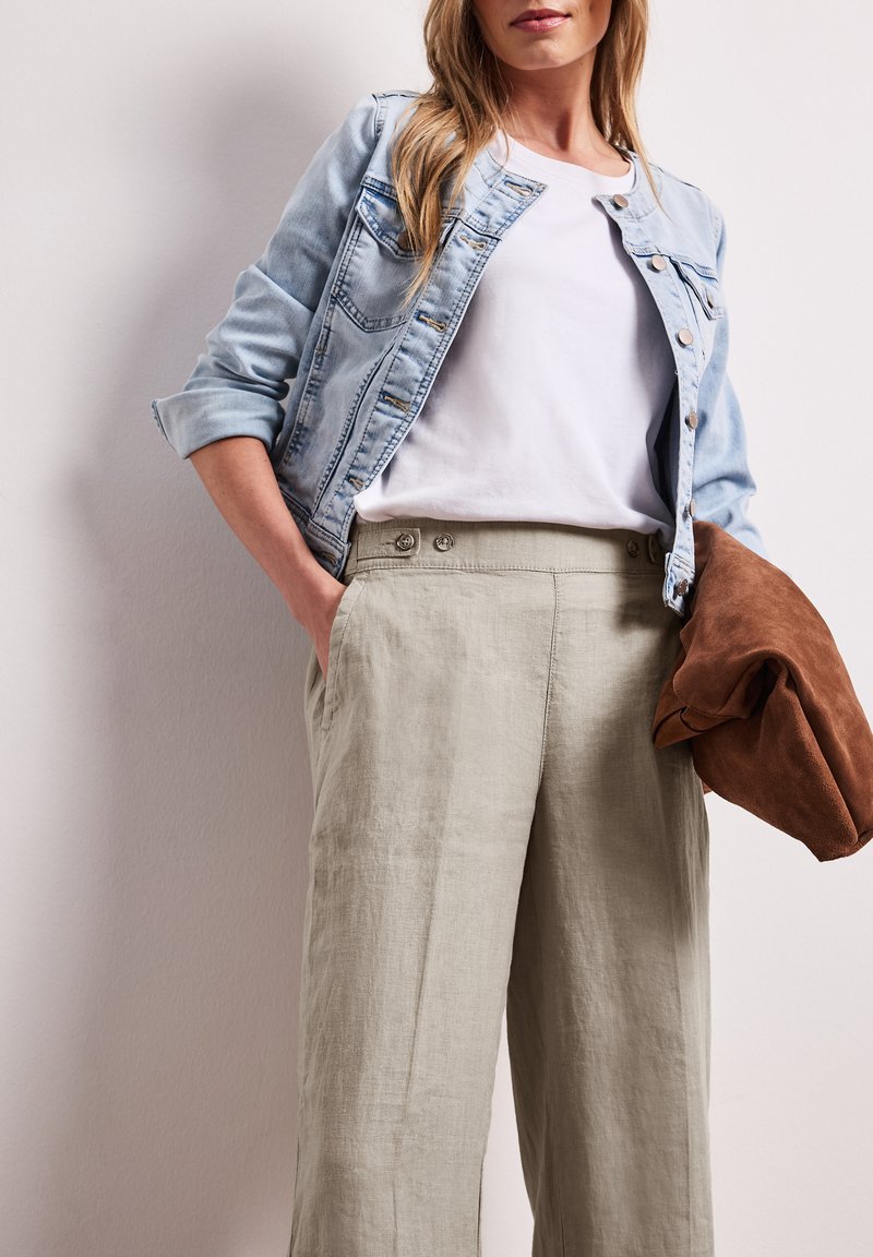 Femme portant une veste en jean claire, une chemise blanche, un pantalon beige taille haute, tenant une veste marron pliée dans une main.