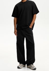 Man med lös svart t-shirt och vida svarta jeans, stående med höger hand i fickan och beige sneakers på vit bakgrund.