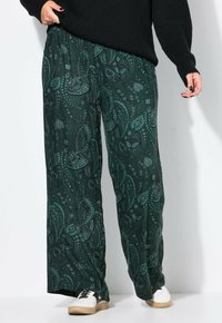 Personne portant un pantalon large en paisley vert foncé, un pull noir et des baskets blanches avec des détails noirs, se tenant devant un fond blanc.