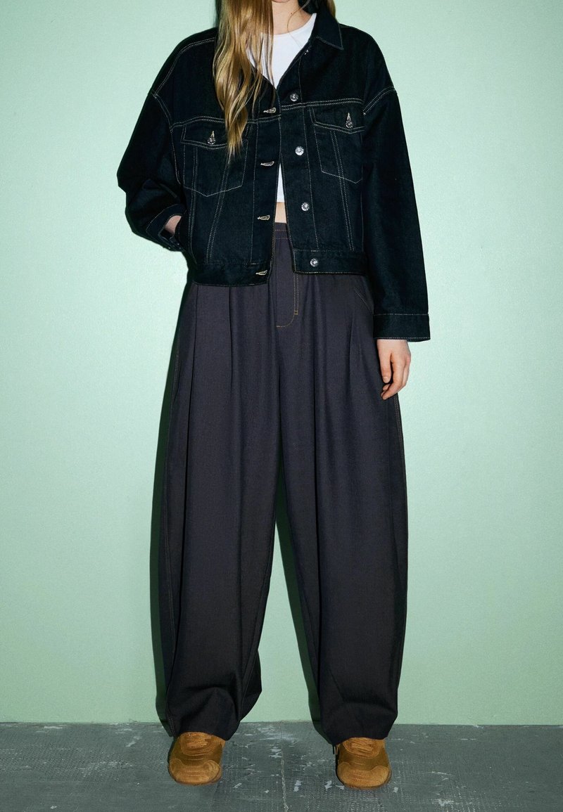 Veste en jean noire avec deux poches avant et boutons en métal, associée à un pantalon large et foncé ainsi qu'à des baskets marron sur un fond vert clair.