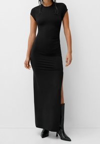 Svart, figursydd maxiklänning i mjukt tyg, med kappramar och en hög slits på låret; kombinerad med knähöga svarta stövlar.