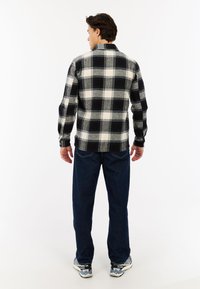Man die met zijn rug naar de camera staat, gekleed in een zwart-wit geruite shirt, donkerblauwe jeans en multicolor sportschoenen op een witte achtergrond.