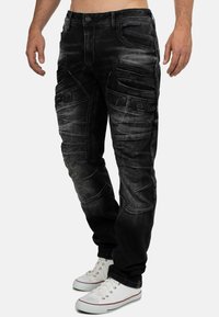 Pantalons cargo en denim noir avec un design délavé, plusieurs poches cargo, des coutures texturées et une coupe décontractée, associés à des baskets blanches.