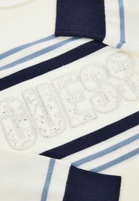 Suéter con base color crema y rayas en azul marino y celeste, adornado con un logo "GUESS" cristalino en detalle brillante.