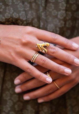 Manos con anillos de oro y perlas, uno con forma de corazón, descansando sobre un fondo de tela floral oscura.